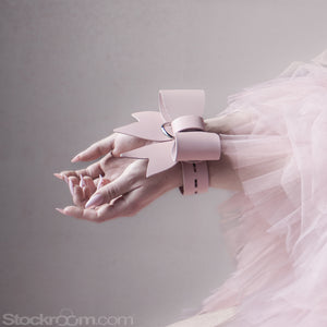 Two feminine hands with long, pink nails extend from the right edge of the picture. They're held together with light pink leather straps, tied in a bow.. 