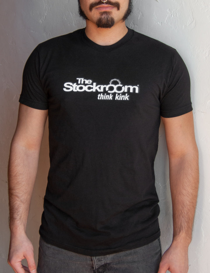 A man is shown wearing a Men's Stockroom Think Kink T-Shirt. The T-shirt is black and has the Stockroom logo printed in white on it, with the slogan “Think Kink” underneath.