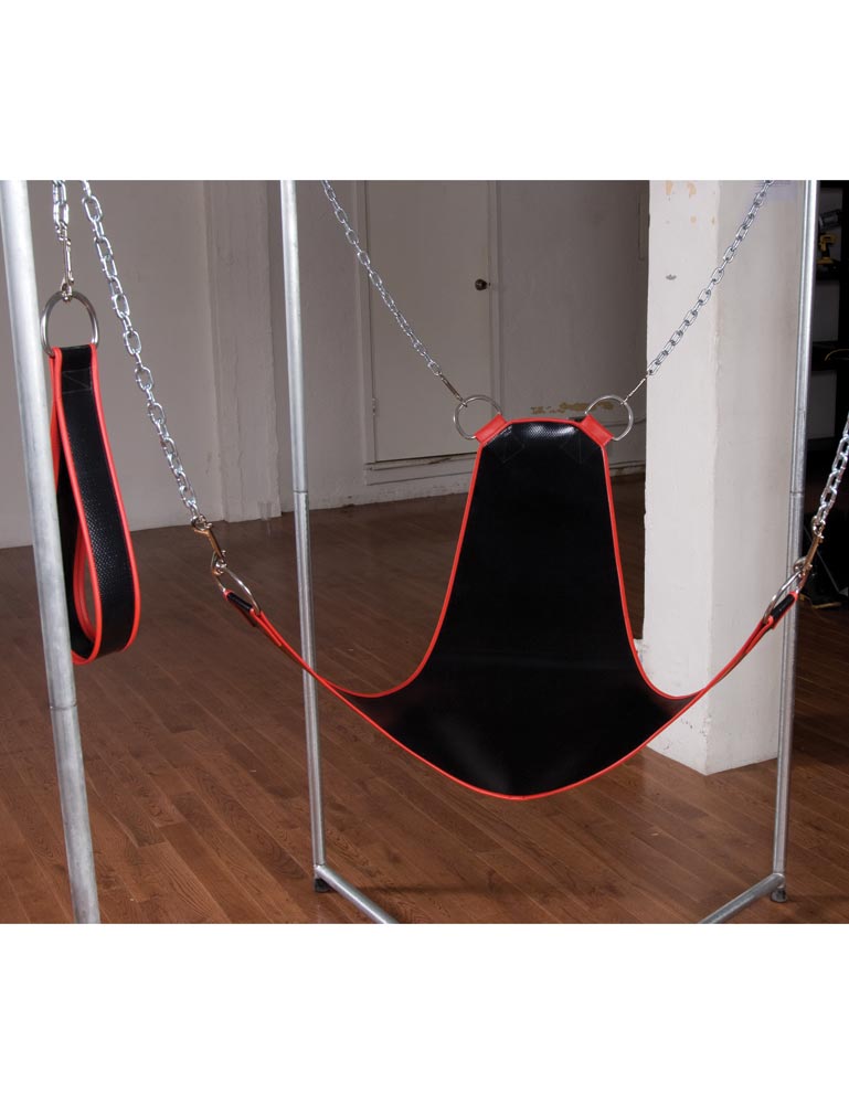 The Pig Sling with Stirrups is shown hanging from a metal structure in a room. The sling is black vinyl with metal trim and has a chain on each corner that is connected to the structure. The chains on the bottom are connected to loop stirrups.