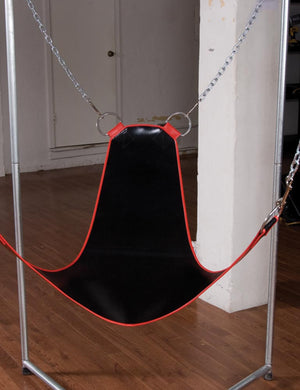 The Pig Sling with Stirrups is shown hanging from a metal structure in a room. The sling is black vinyl with metal trim and has a chain on each corner that is connected to the structure. 
