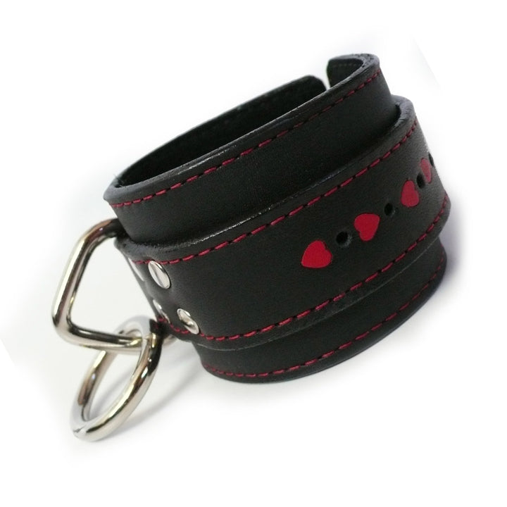 A single Wrist Restraint With Heart Inlay is displayed against a blank background. The cuff is made of black leather with red accent stitching. Around the middle of the cuff, heart-shaped pieces are cut out, revealing red leather underneath.