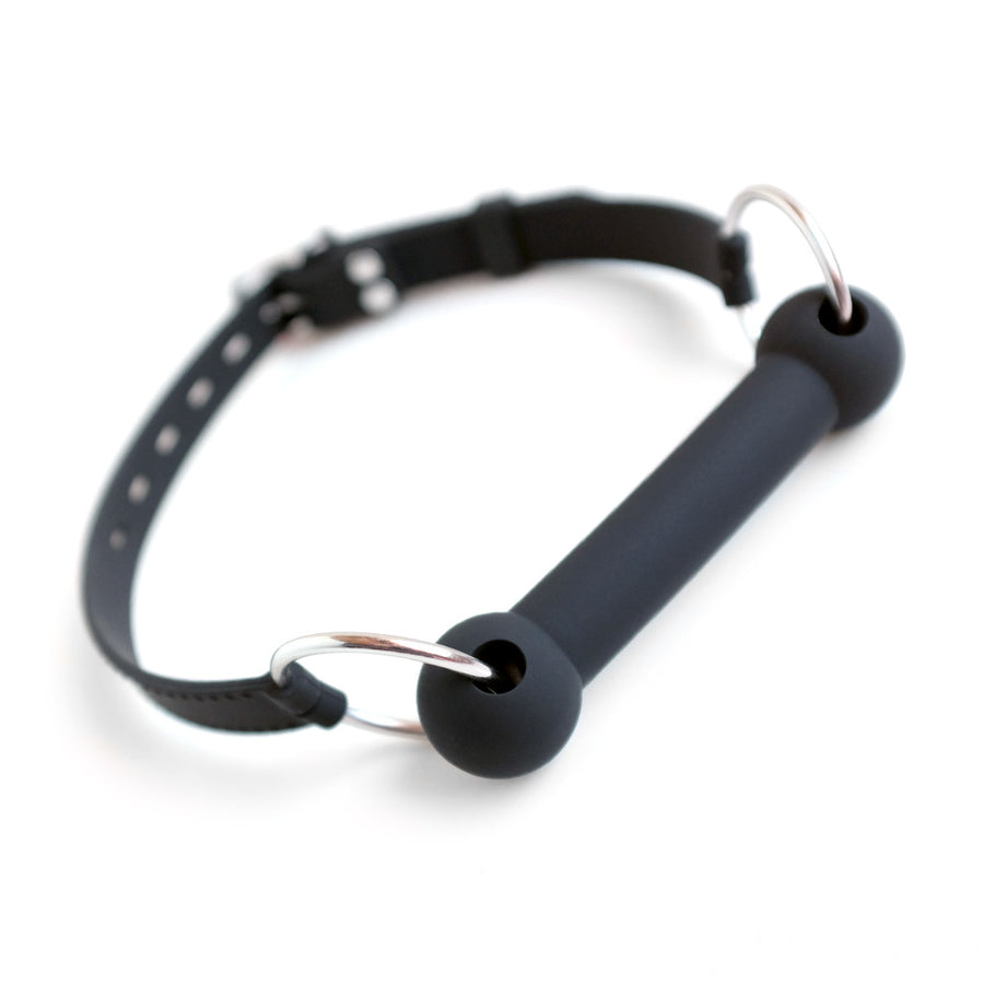 The Silicone Bit Gag With Silicone Strap is displayed against a blank background. The gag is made of matte black silicone and is shaped like a bit. Each end of the gag has two metal O-rings that connect to the strap.