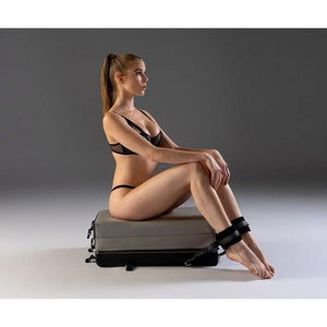 A woman in black lingerie with her hair in a ponytail is sitting on a folded-up Bondi Portable Playscape By Liberator. She is wearing ankle cuffs, which are attached to the Liberator.