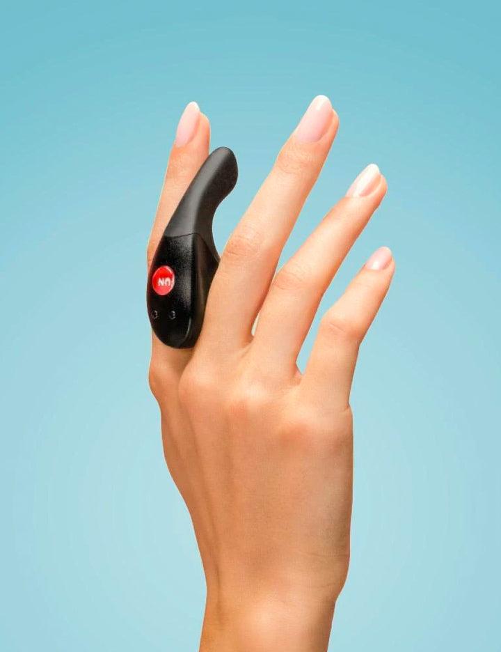 A person’s hand is shown holding the Fun Factory Be-One Couples Vibrator between their fingers against a light blue background.