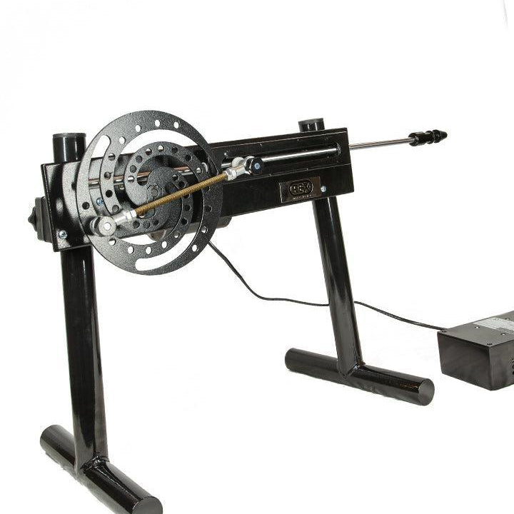The Rex Machine is displayed against a blank background. It is a sex machine made of black metal. It is elevated on two legs and has a spiral wheel at the back with a gear attached leading to a pole. There is a grey control box next to it. 