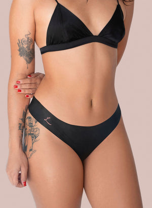 A woman in a black bra and bikini style Lorals Dental Dam Latex Panties is shown standing against a pink background. The panties are black with a small, pink “L” on one hip.
