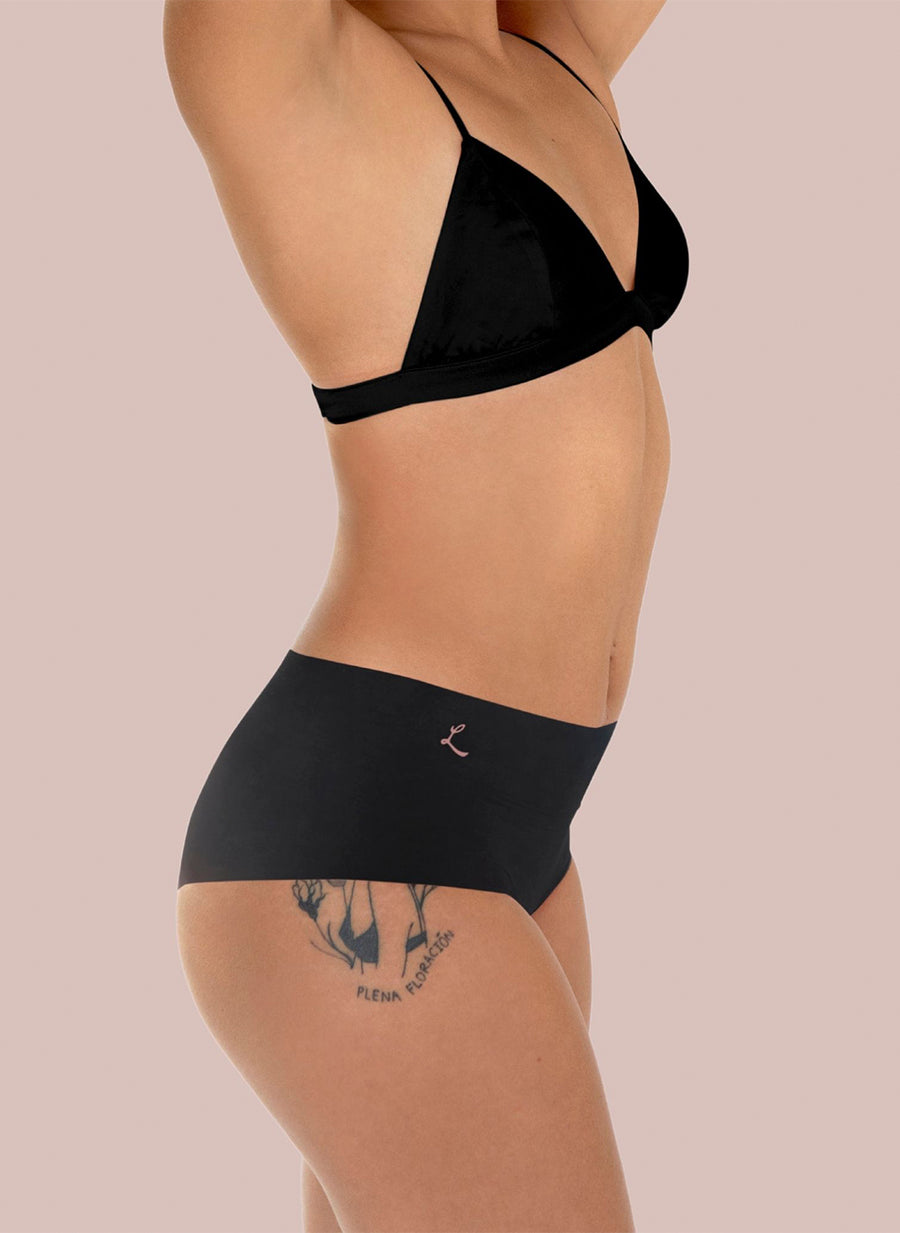 A woman wearing black bra and bikini style Lorals Dental Dam Latex Panties is shown standing in profile against a pink background with her arms above her head.