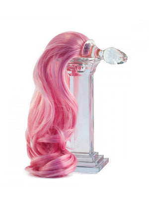 The Pink Detachable Ponytail Glass Butt Plug rests ontop of a clear pedestal in front of a blank background. The tail is draped down the side of the pedestal.