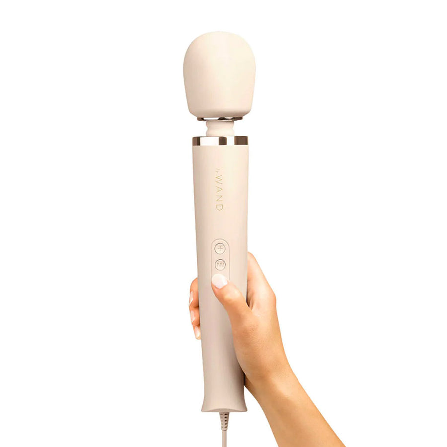 A woman’s hand with white nail polish is shown holding up a Le Wand Plug-In Vibrating Massager in Cream against a blank background.