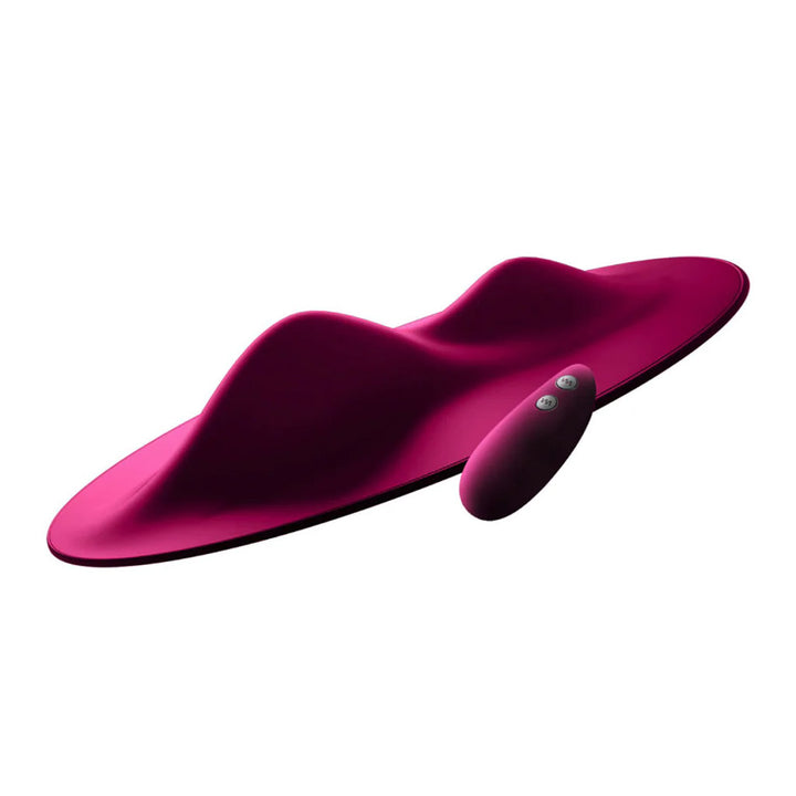 The Vibepad Remote Controlled Grinding Vibrator Pad is shown against a blank backgroud. The toy is dark pink and is shaped like a disc with 2 prominent mounds, one small and one large. Its remote, which has 2 buttons, is shown next to it.