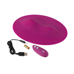 The Vibepad Remote Controlled Grinding Vibrator Pad and the contents of its package are shown against a blank backgroud. The toy is shown alongside its charger, its remote, and an A23 battery for the remote.