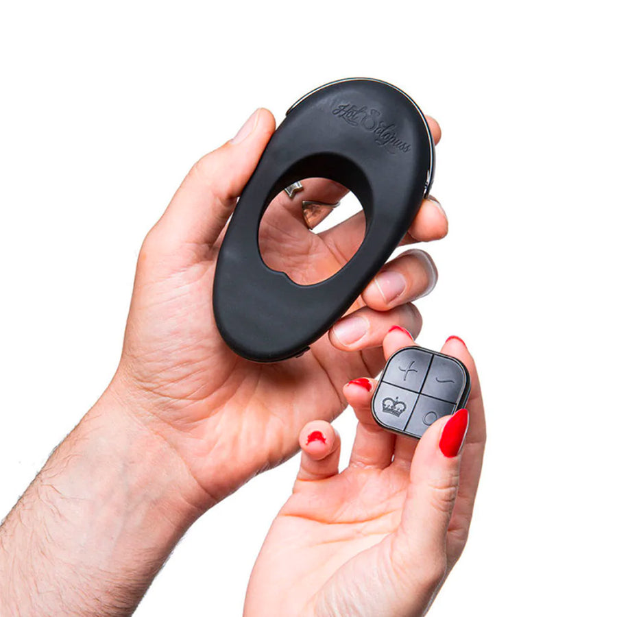 A man’s hand is shown holding up the The Hot Octopuss Atom Plus Lux Vibrating Cock Ring while a woman holds up the remote. The remote is square and has four buttons.