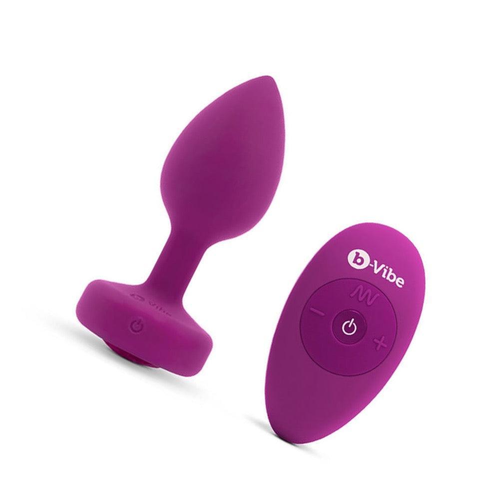 The b-Vibe Vibrating Jewel Butt Plug is shown against a blank background next to its 4 buttoned remote. The plug is a size S/M, which is Fuchsia colored. It is tapered with a thin neck and flared base.