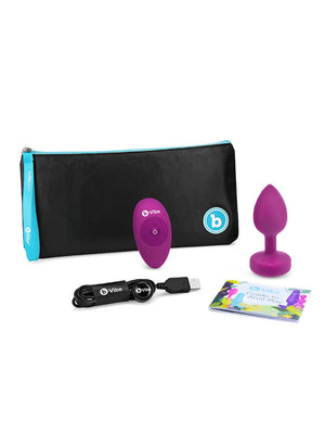 The contents of the packaging for the b-Vibe Vibrating Jewel Butt Plug in fuscia are shown against a blank background. Displayed are the plug, the remote, the charger, a guide to anal play, and a black cloth pouch with a blue zipper.