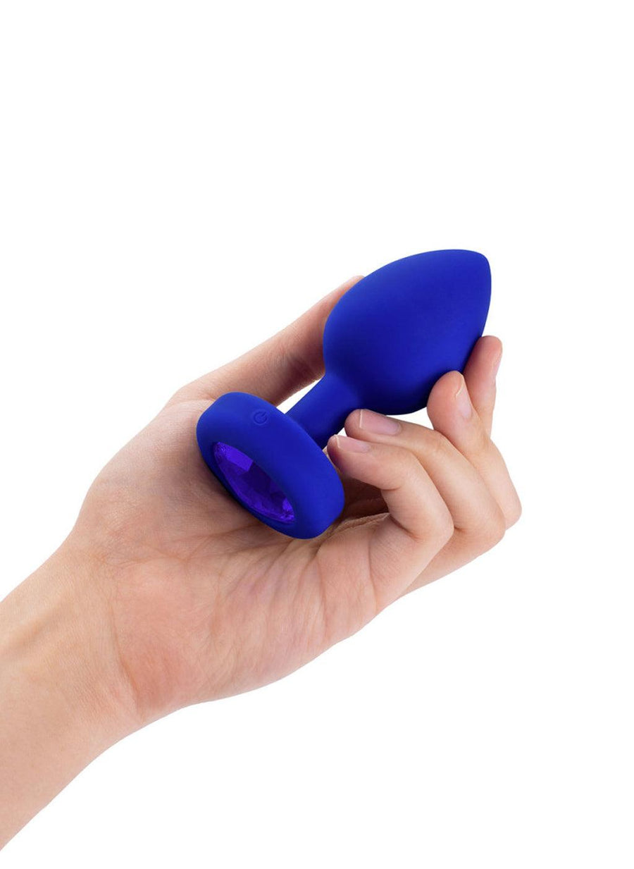 A hand is shown holding the b-Vibe Vibrating Jewel Butt Plug in Blue Sapphire against a blank background on its side, displaying the blue crystal on the base.
