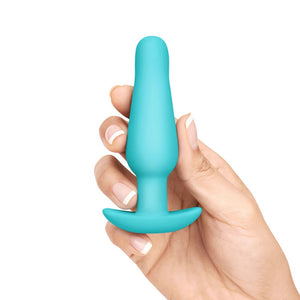 A hand with french-tip nails is shown holding up the smallest plug from the b-Vibe Anal Training & Education Butt Plug Set in Aqua against a blank background.