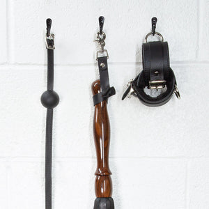 A close-up image of the Sissoo Rosewood Long Handle Leather Flogger with leather hanging strap on a white background. The flogger is shown hanging on a black hook next to leather wrist cuffs and a black silicone ball gag made by The Stockroom. 
