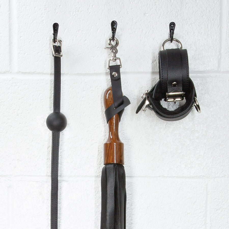 A close-up image of the Sissoo Rosewood Short Handle Leather Flogger with leather hanging strap on a white background. The flogger is shown hanging on a black hook next to leather wrist cuffs and a black silicone ball gag made by The Stockroom. 