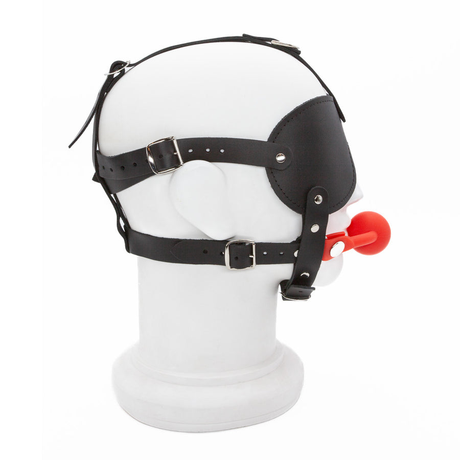 The Silicone Ball Gag And Blindfold Head Harness is shown on a mannequin head from the side. The red silicone ball gag connects to the chin straps.