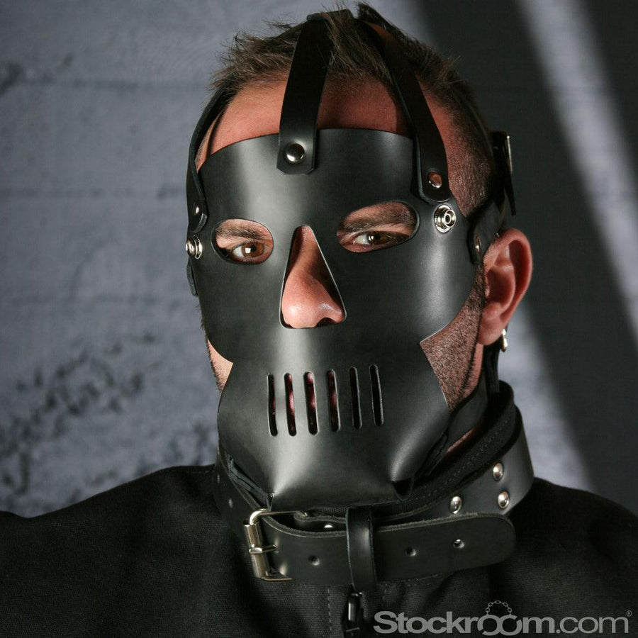 A close-up of a man wearing The Punisher Muzzle Leather Head Harness is shown. The harness covers his face with holes for the eyes and nose and vertical slits over the mouth.