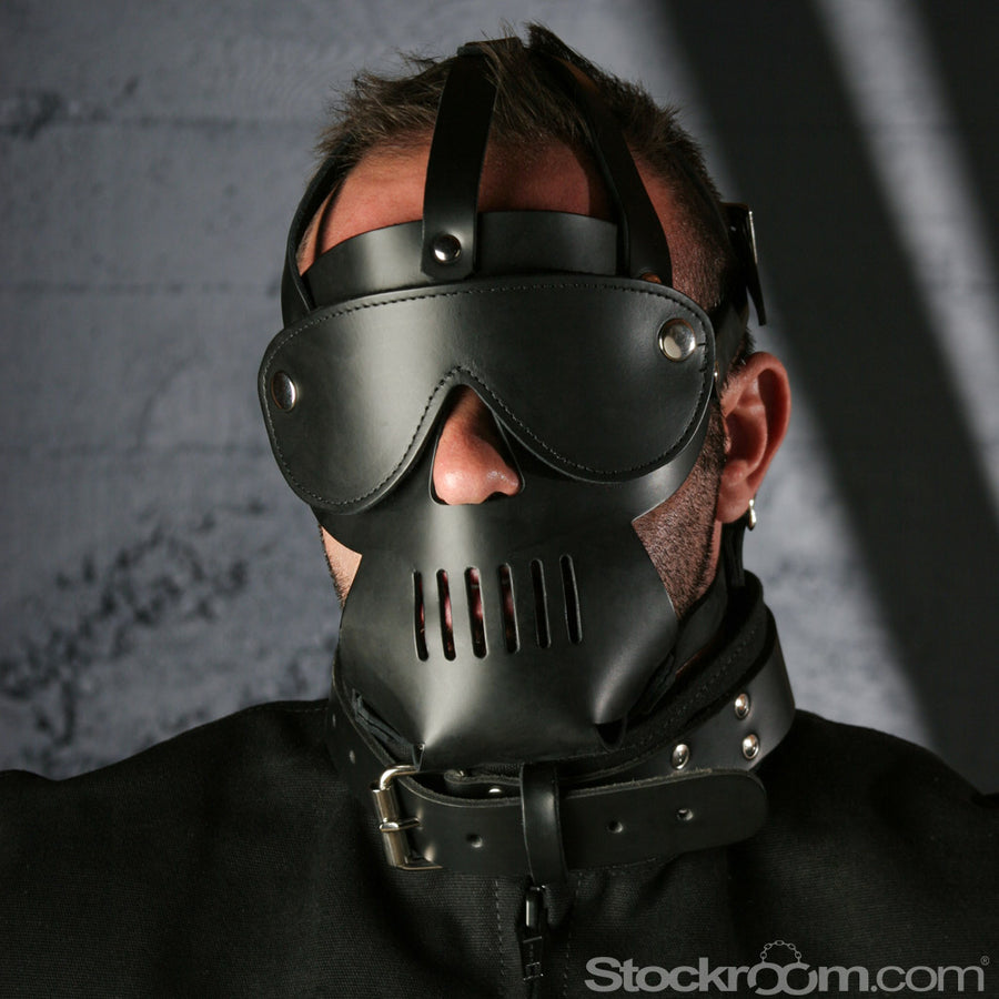 A close-up of a man's head in The Punisher Muzzle Leather Head Harness is shown. The hood is made of black leather and covers the mouth with a skull design. A blindfold is snapped on to the harness.