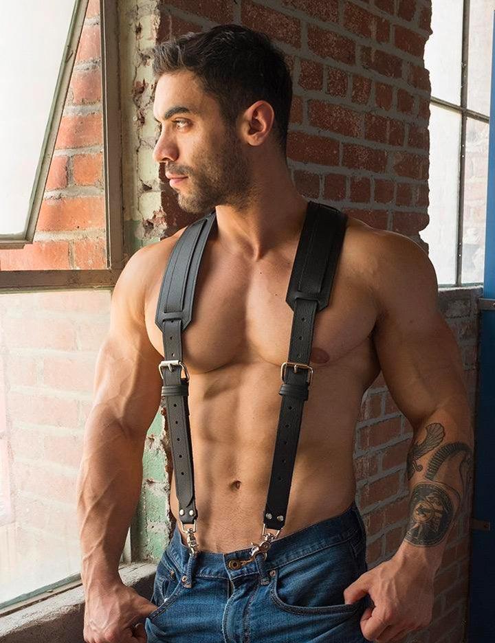 A shirtless, muscular man with facial stubble stands looking out a window. He wears blue jeans and black Deluxe Leather Suspenders that clip to his belt loops. The suspenders have buckles in the front and wider pieces of leather on the shoulders.