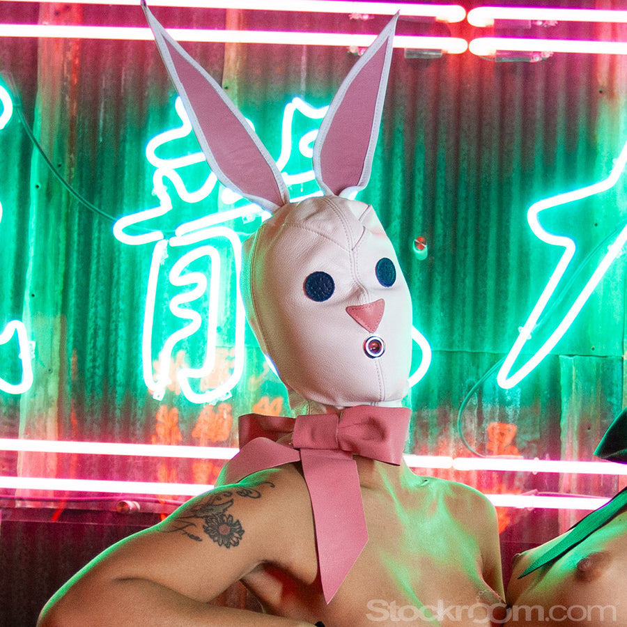 A nude woman reclines in front of a green and red neon sign. She wears the white Leather Bunny Hood, which covers her face and head and ties with a pink bow around her neck. The hood has large ears, blue eyes, a pink nose, and a small opening over the mouth.