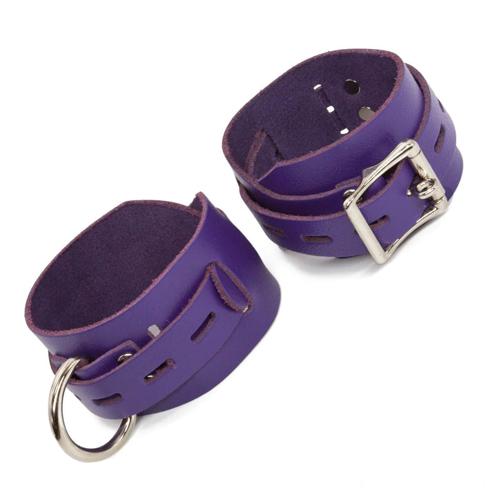A pair of purple Locking/Buckling Leather Bondage Restraints are displayed against a blank background. They are made of a wide piece of leather with a narrower, notched piece of leather wrapping around them. Each cuff has a silver D-ring and a lockable buckle.