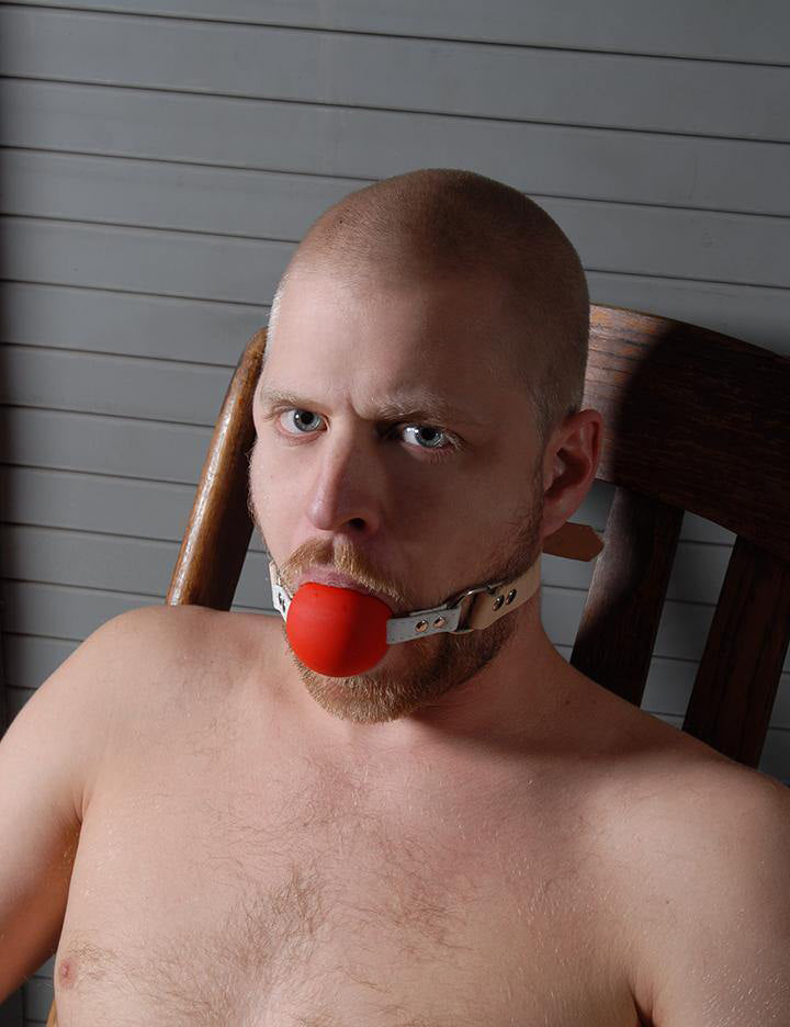 A shirtless man with a buzzed head and facial hair is shown sitting in a wooden chair. He has the Medical Silicone Ball Gag in his mouth.