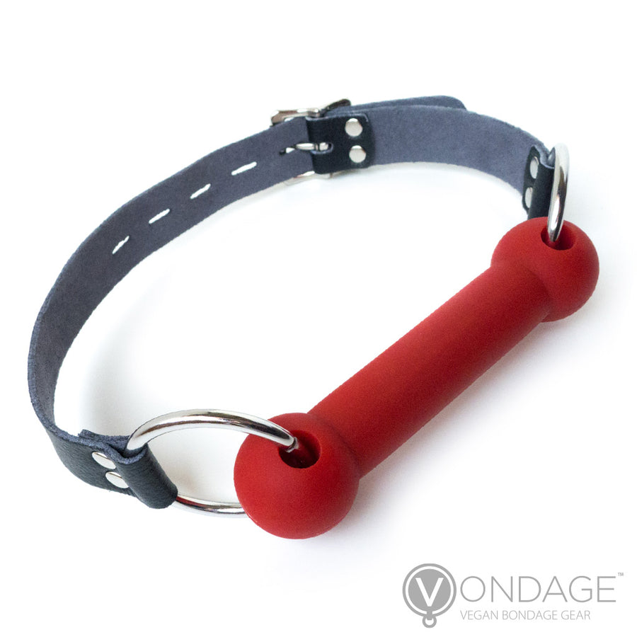 The Vondage Silicone Bit Gag is shown against a blank background. The gag is made of matte red silicone and shaped like a bit with metal O-rings on the sides that connect it to the adjustable vegan leather strap.