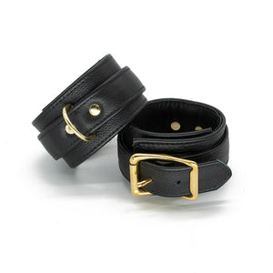 A pair of black Garment Leather Ankle Cuffs With Brass Gold Hardware are shown buckled against a blank background. 