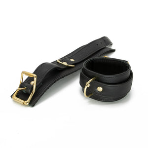 A pair of black Garment Leather Ankle Cuffs With Brass Gold Hardware are shown against a blank background. One of the cuffs is buckled and one is laying flat.