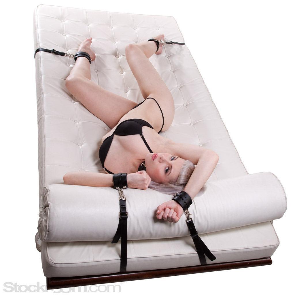 A woman in black lingerie lays on her side on a white chaise lounge. Her arms are above her head, and she wears black wrist and ankle cuffs. 
