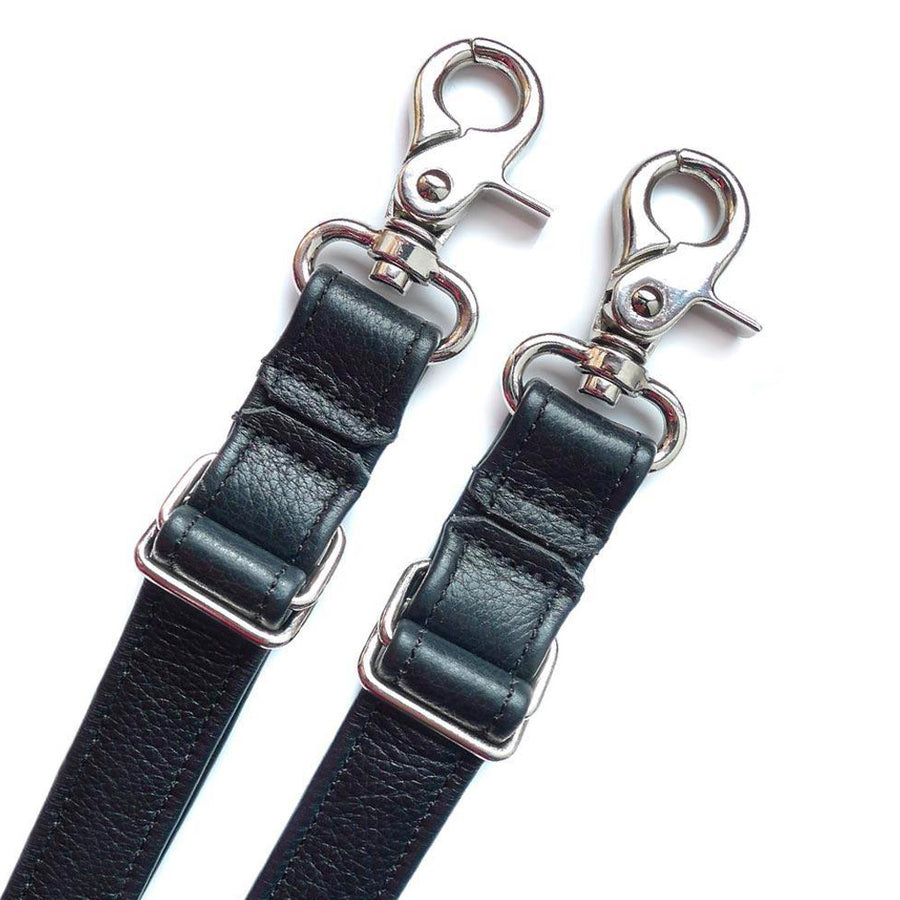 A close-up of the ends of two of the restraint straps from the Kinklab Bound-O-Round 4-Point Restraint System is shown against a blank background. The straps are made of black leather and have silver metal snap hooks on the ends.