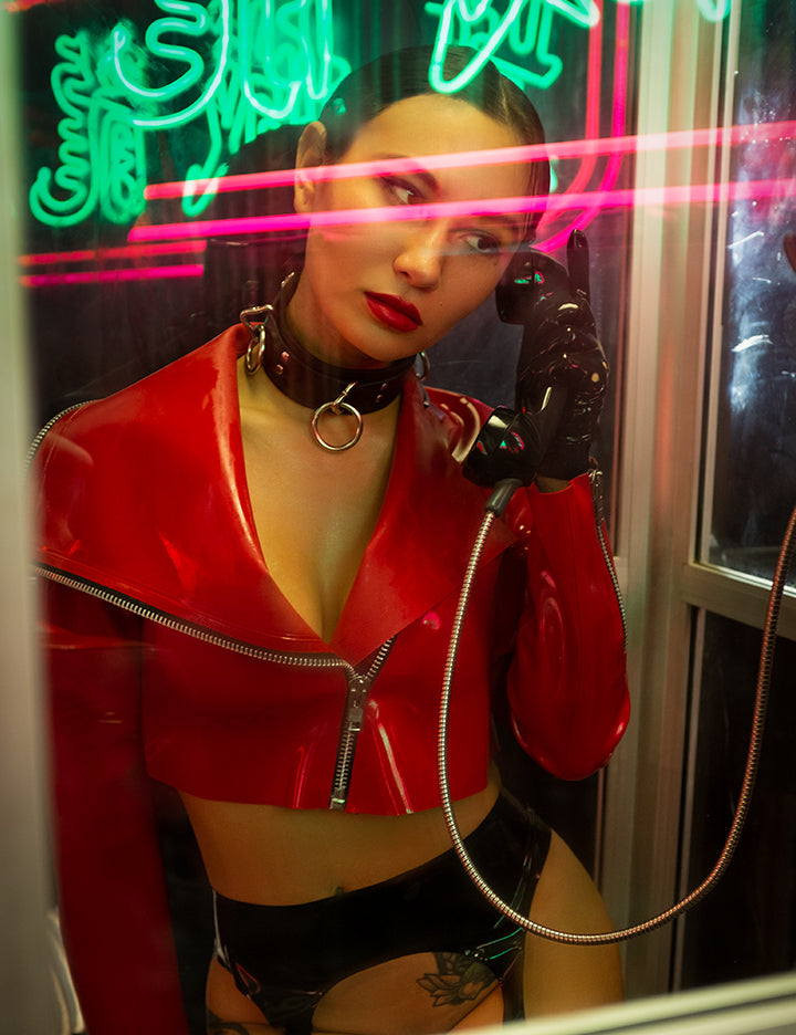 A woman with her hair tightly pulled back sits in a phone booth, holding the phone to her ear, with neon lights reflected on the glass. She wears the Zeta Jacket by Syren Latex in red, black latex gloves, and a large black collar. 