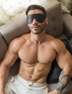 A muscular brunette man in white sweatpants reclines on a grey couch. He is wearing the Padded Leather Blindfold.