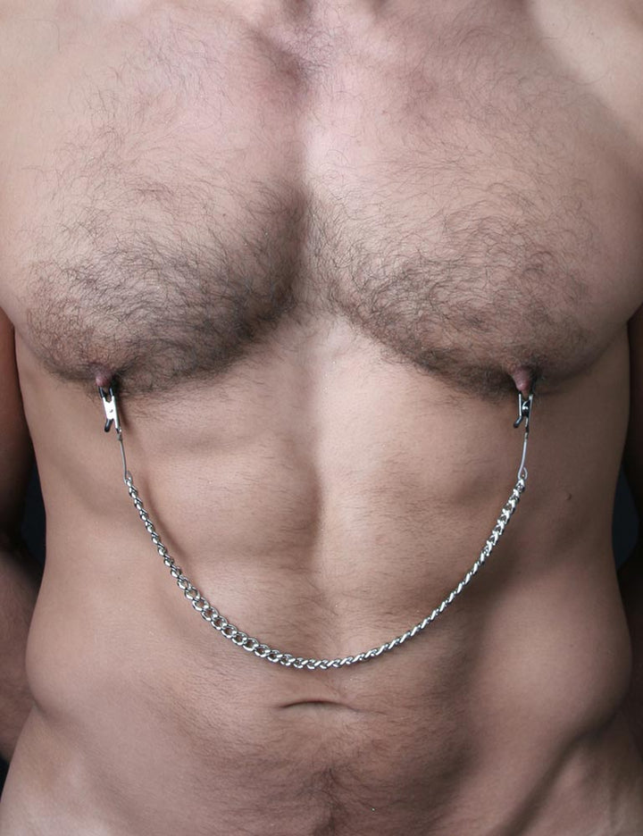 A close-up of a muscular man’s bare torso with some body hair is shown. He wears the silver Mini Nipple Clamps.
