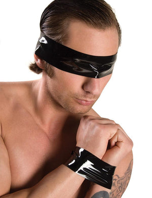 A shirtless brunette man is shown from the chest up in front of a blank background with his arms held in front of his chest. Black bondage tape binds his wrists together and covers his eyes.