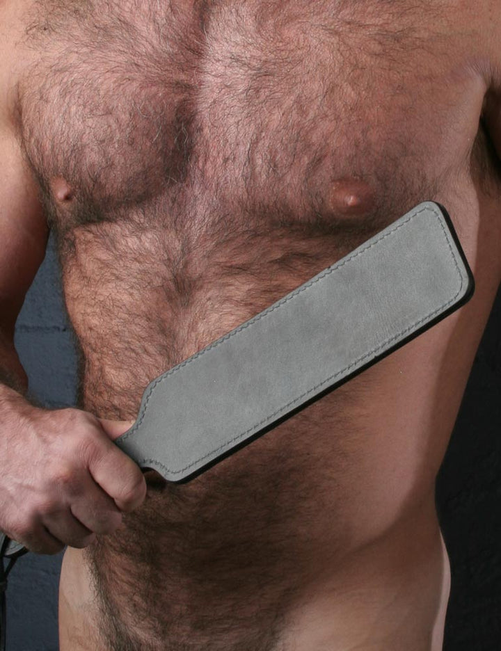 A close-up of a nude man’s hairy chest is shown. He holds the Black Leather Fraternity Paddle in his fist in front of his chest.