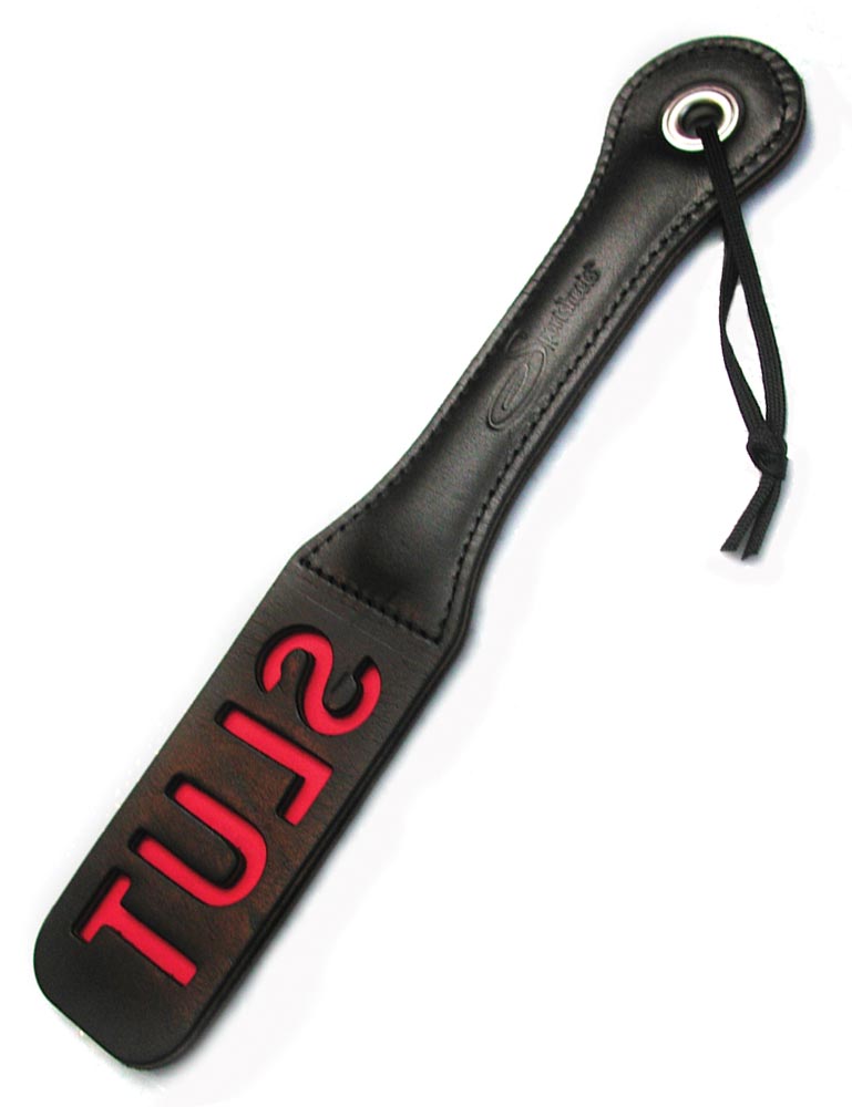 The Slut Impression Paddle is displayed against a blank background. The paddle is rectangular with a wrist loop and made of black leather with the word slut cut out, revealing red leather underneath.