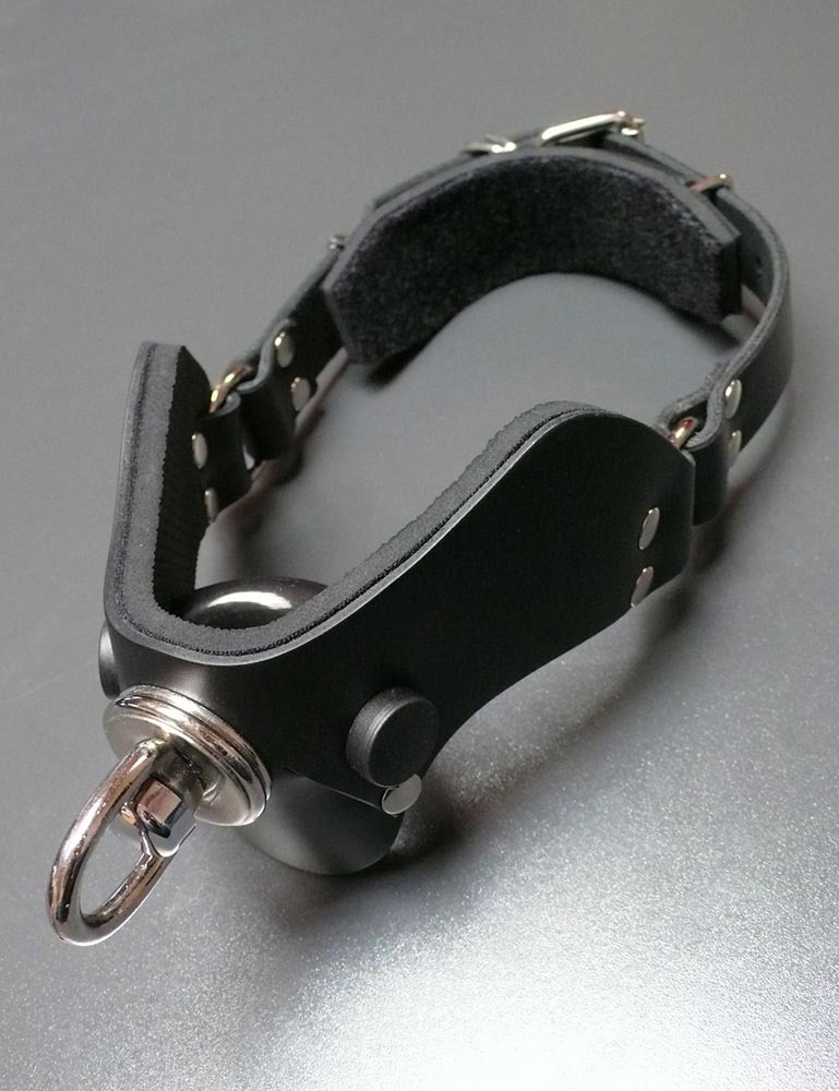 The Half Moon Bit Gag By Scott Paul Designs is displayed on a grey table. The exterior of the gag is lined with black leather, and the interior has black padding on the front of the gag and behind the buckle.