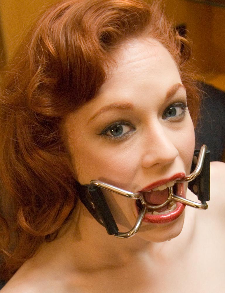 A close-up of a red-haired woman shows her wearing The Spider Gag. The gag's metal O-ring is in her mouth, and metal brackets attached to it keep her mouth open and attach to black leather straps.