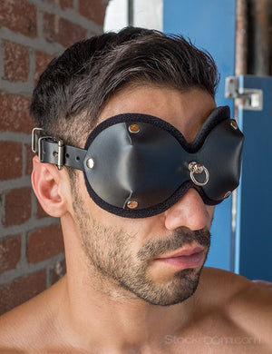  A close-up of a brunette man with light facial hair is shown. He wears the black Mistress Heather Leather Blindfold, which has cupped eye covers with a small metal D-ring between the eyes.
