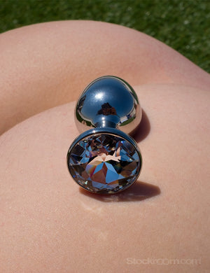 A close-up of a person’s bare ass is shown with green grass in the background. The crystal Julian Snelling Rosebuds Jeweled Anal Plug With A Swarovski Crystal rests on top of their ass. The plug is silver metal, and the base is covered by a clear crystal.
