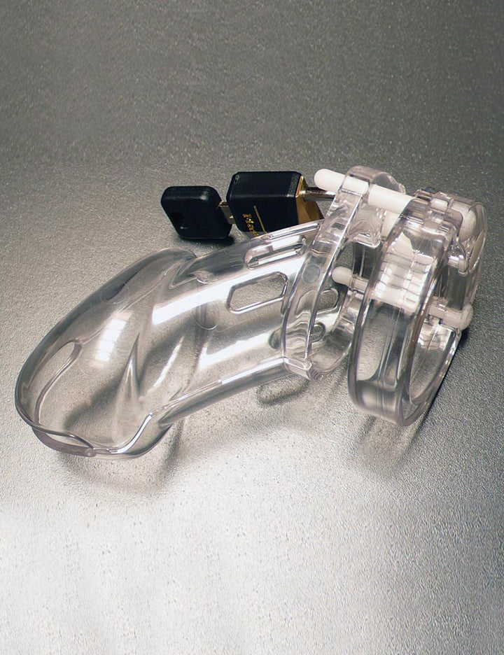 The assembled cage from the CB-6000 Package Male Chastity Kit is shown against a silver background. The cage is made of clear plastic with small cutouts and is shaped like a flaccid penis. It is attached to the base ring and locked with a small padlock.