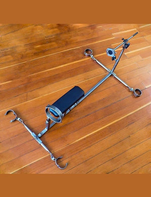 The Stockroom Stockade is shown on a wooden floor. It has 1 vertical bar with 2 horizontal spreader bars with metal cuffs. It has an upright bar with a metal collar and a fucking rod at the other end. The leather stomach support is not upright.