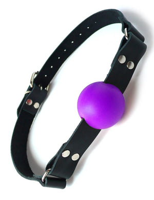 The purple rubber ball gag is displayed against a blank background. It is a matte purple rubber ball attached to a black leather strap with silver hardware, which buckles behind the head.
