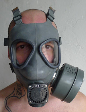 A close-up of a man with buzzed hair standing in front of a grey wall wearing the grey Finnish Military Gas Mask with the side-mounting filter attached. 