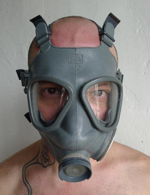 A close-up of a man with buzzed hair standing in front of a grey wall wearing the grey Finnish Military Gas Mask with the filters removed.