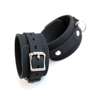 The Silicone Bondage Locking Ankle Cuffs are displayed against a blank background. They are made of matte black silicone with faux stitching along the borders. They are adjustable and have a lockable buckle and one D-ring on each cuff.